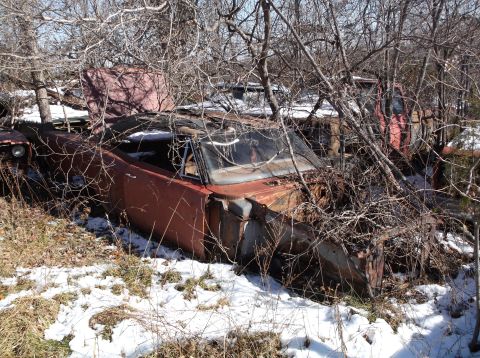 1974 Dodge Challenger - A rusty, red convertible car, likely a 1974 Dodge Challenger, sits abandoned in a wooded area with snow on the ground. The vehicle is heavi…