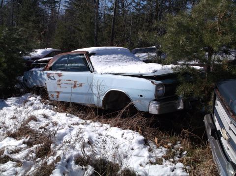 1974 Dodge Challenger - A light blue coupe sits in a snowy junkyard, its hood and roof covered in snow, with other vehicles visible in the background.