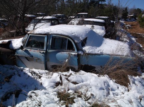 1974 Dodge Challenger - A forgotten blue and white sedan sits buried in snow amongst other vehicles in a junkyard.