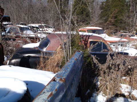1974 Dodge Challenger - A collection of vintage vehicles, mostly cars and trucks, sit in a snowy junkyard setting surrounded by trees. The vehicles are heavily rus…