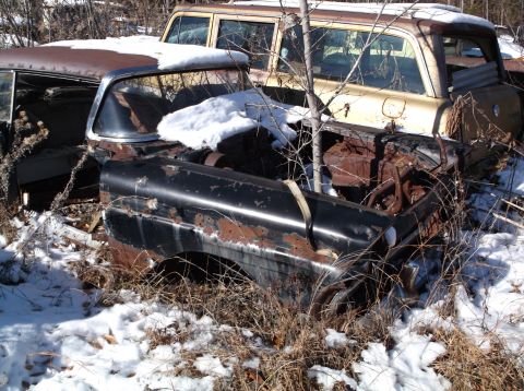 1974 Dodge Challenger - Two heavily rusted and snow-covered vintage vehicles are parked outdoors in a wooded area. The front vehicle is a black sedan, and behind i…