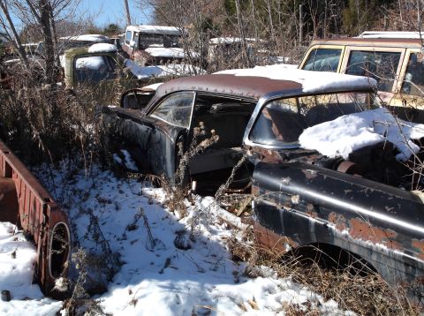 1974 Dodge Challenger - A rusty, black coupe sits in a junkyard surrounded by snow and dry brush. The driver's side door is open, revealing a dark interior.