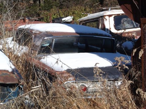 1974 Dodge Challenger - A rusty, snow-covered vintage sedan is partially obscured by dry weeds and branches. Another older vehicle, possibly a van or truck, is vis…