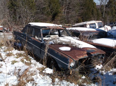 1974 Dodge Challenger - A rusty, dark blue sedan sits in a snowy junkyard, partially covered in snow and dry vegetation. The front end of the car is heavily damage…