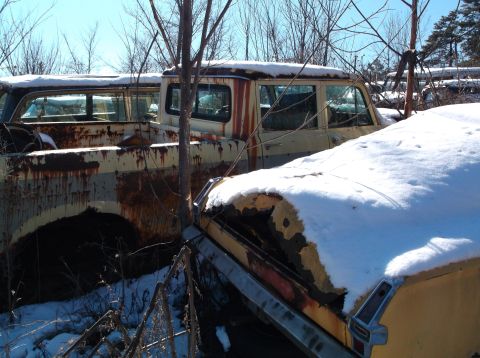 1974 Dodge Challenger - Two heavily rusted and snow-covered vintage vehicles, likely trucks or vans, are parked outdoors in a junkyard setting with bare trees and…