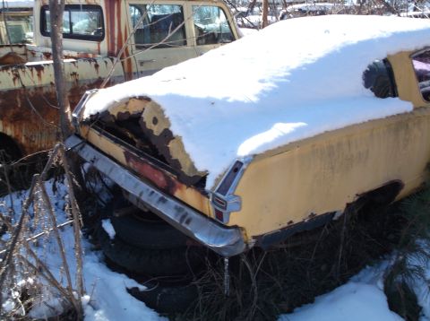 1974 Dodge Challenger - A rusty, yellow vintage car, partially covered in snow, sits in an outdoor junkyard setting. The rear of the vehicle shows significant rust…