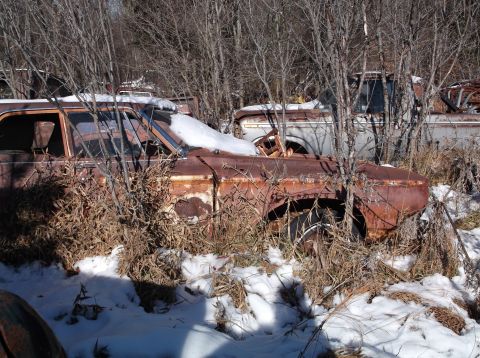 1974 Dodge Challenger - A heavily rusted vintage car, possibly a 1974 Dodge Challenger, sits in a snowy junkyard surrounded by bare trees and dry brush. Another ve…