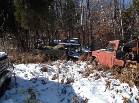1974 Dodge Challenger - Two abandoned vehicles, a green coupe and a red truck, sit in a snowy wooded area. The coupe is heavily rusted and missing parts, while the…