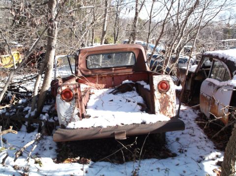 1974 Dodge Challenger - The rear of a rusty, abandoned pickup truck covered in snow sits in a wooded area. The truck has a red cab and bed, with round taillights o…