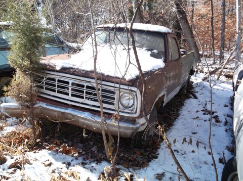 1974 Dodge Challenger - An abandoned, rusty Dodge pickup truck sits in a snowy wooded area, partially covered in snow. The front grille and headlight are visible.