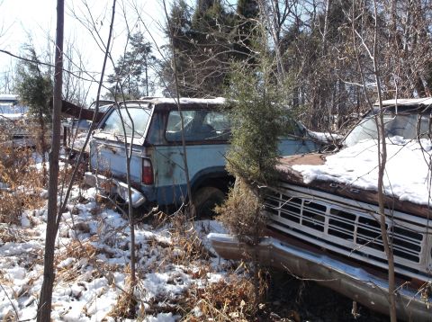 1974 Dodge Challenger - Two old, rusty pickup trucks are parked in a wooded area with snow on the ground. The truck in the foreground is a reddish-brown color and…