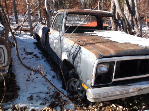 1974 Dodge Challenger - A rusty, dark gray pickup truck sits in a wooded area with snow on the ground and on its hood. The front of the truck is visible, showing a…