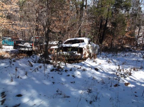 1974 Dodge Challenger - A rusted and snow-covered car body, likely a coupe, sits abandoned in a wooded area with other vehicles visible in the background.