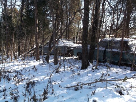 1974 Dodge Challenger - Two vintage teal-colored sedans are partially covered in snow and surrounded by trees in a wooded area. The vehicles appear to be abandoned…