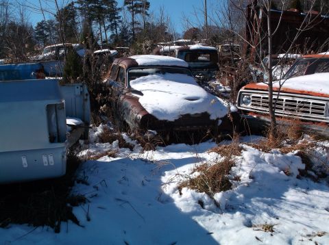 1974 Dodge Challenger - Several old, snow-covered vehicles are parked in a junkyard on a sunny day. A rusty sedan with snow on its hood is in the foreground, with…