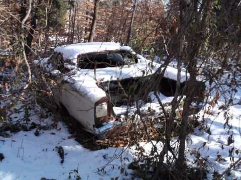 1974 Dodge Challenger - An abandoned white car, likely a coupe, is covered in snow and surrounded by trees in a wooded area. The vehicle appears to be in a state o…