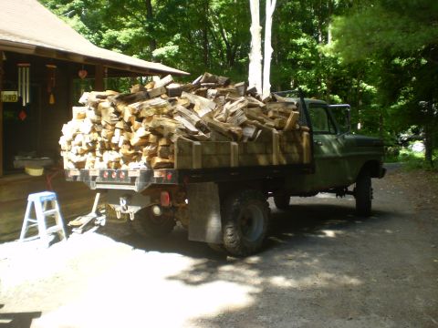 1979 Ford Bronco - A green flatbed truck is loaded with a large pile of chopped firewood, parked next to a rustic wooden house with a porch. A small step ladd…