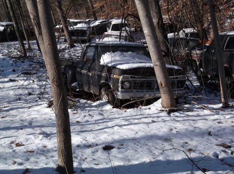 1974 Dodge Challenger - A black pickup truck covered in snow sits in a wooded area amongst other vehicles. The truck appears to be an older model with visible rust.