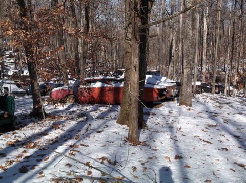 1974 Dodge Challenger - A red convertible sits abandoned in a snowy forest junkyard, its front end showing signs of disrepair.