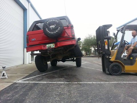 1995 Ford Bronco - A red Ford Bronco is being lifted by a forklift. The spare tire cover clearly shows 'Ford Bronco'.