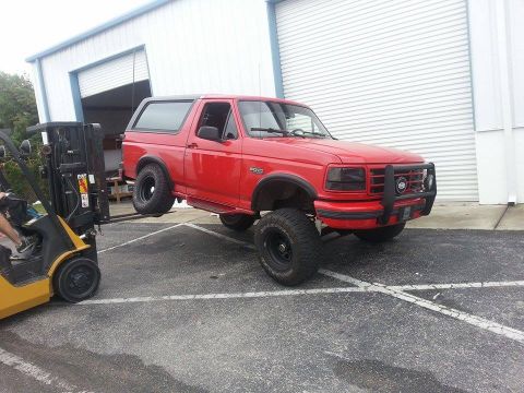 1995 Ford Bronco - A red Ford Bronco is being lifted by a forklift outside a building with a large garage door.