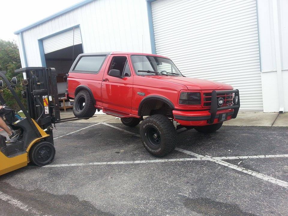 Red Ford Bronco with aftermarket bull bar and large tires, lifted by a yellow forklift