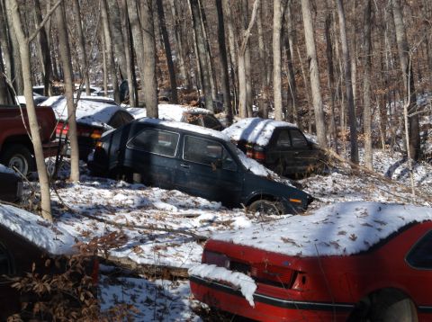 1974 Dodge Challenger - Multiple vehicles are seen in a wooded area, covered in snow, suggesting a junkyard setting.