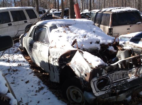 1974 Dodge Challenger - A snow-covered 1974 Dodge Challenger sits in a junkyard, showing significant rust and damage.
