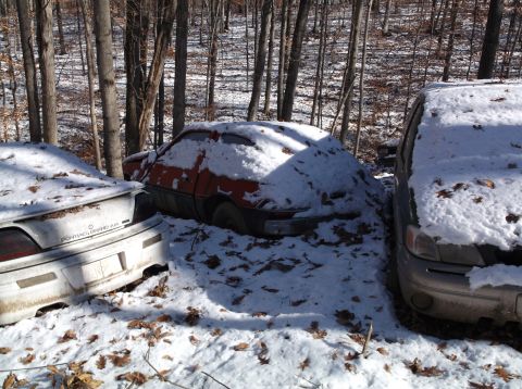 1974 Dodge Challenger - Several vehicles sit abandoned and covered in snow amongst trees in a wooded area.