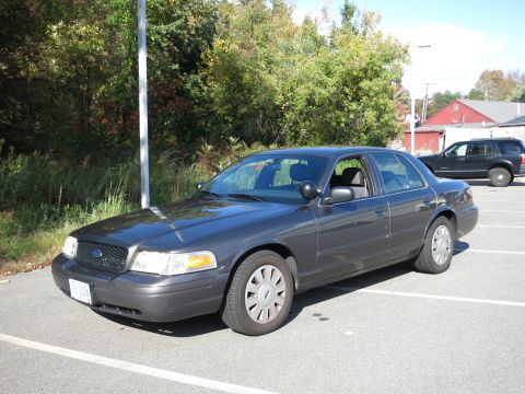2001 Ford Crown Victoria - A dark grey Ford Crown Victoria is parked in a lot on a sunny day, with trees and a building in the background.