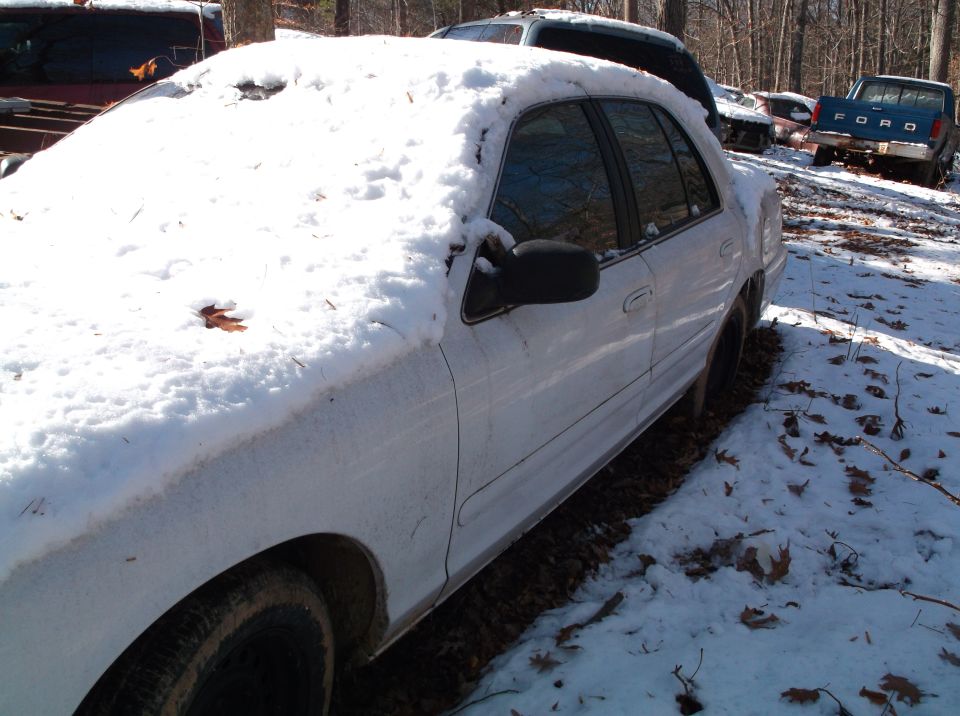 White sedan partially covered in snow, parked in a wooded area with other vehicles visible in the background