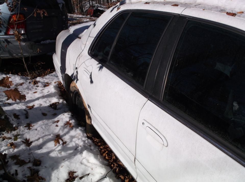 White sedan parked in snow, next to a dark SUV. Some leaves on the ground