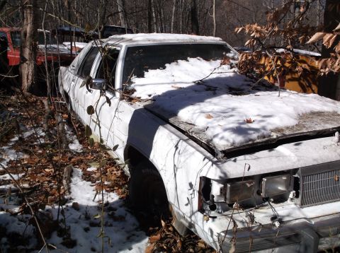 1974 Dodge Challenger - A white sedan sits abandoned in a snowy junkyard, partially covered by snow and fallen leaves.