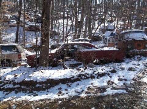 1974 Dodge Challenger - Multiple abandoned vehicles sit in a snowy, wooded junkyard, including a red muscle car.