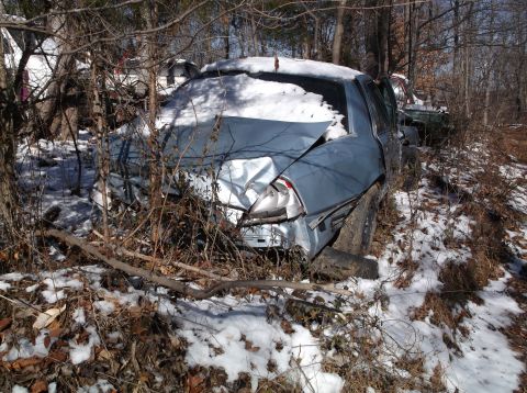 1974 Dodge Challenger - A damaged blue car sits abandoned in a snowy junkyard, partially obscured by trees and debris.
