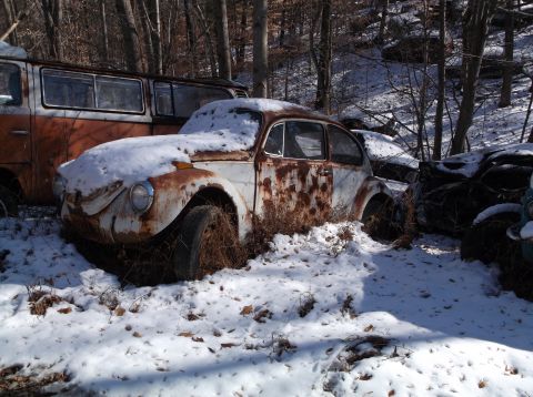 1974 Dodge Challenger - A rusty Volkswagen Beetle sits abandoned in the snow, surrounded by other vehicles in a forest.