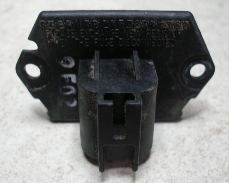 Black blower motor resistor with two mounting holes and a plastic connector. Text is visible on the black housing