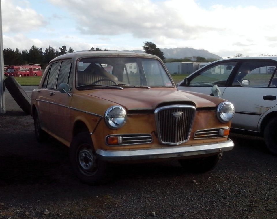 Old orange sedan parked next to a white car. Tire leaning against a pole on the left