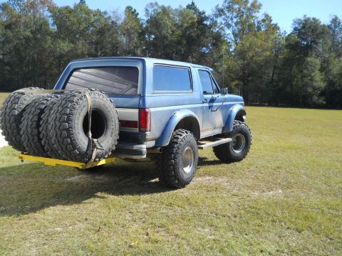 1992 Ford Bronco - A blue Ford Bronco is parked in a grassy field with three large tires mounted on a yellow rack on the rear. The vehicle has been modified w…