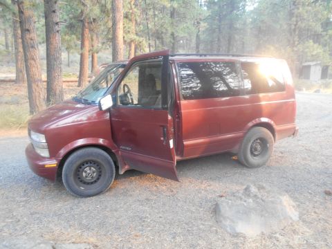 2000 Chevrolet Astro - A maroon Chevrolet Astro van with the driver's side door open is parked on a gravel road in a wooded area. The van has tinted windows and a…