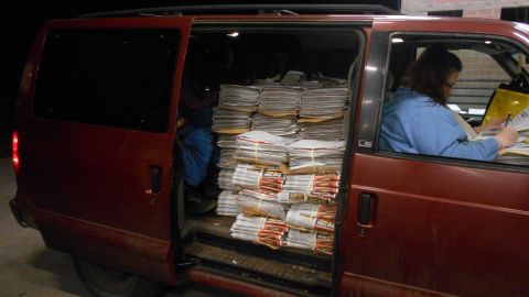 2000 Chevrolet Astro - The rear cargo area of a maroon minivan is packed with stacks of newspapers. A woman is visible in the driver's seat, looking at papers.