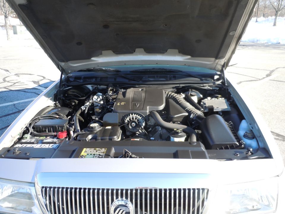 Silver sedan engine bay with V8 engine, battery, air intake, and alternator visible.