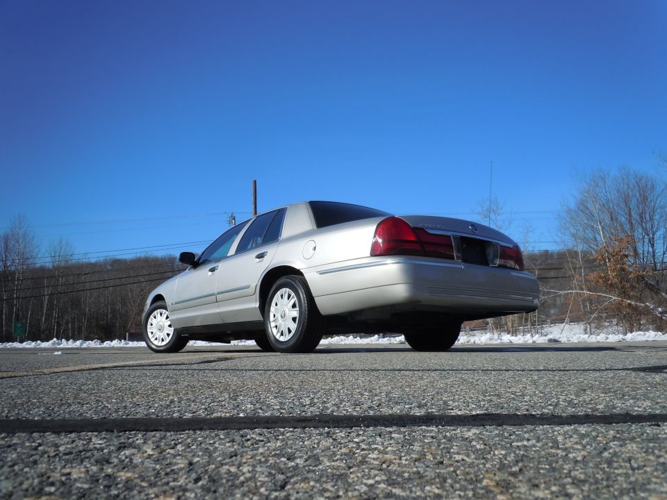 Silver 2001 Ford Crown Victoria, rear passenger side view, parked on a road with snow and trees.