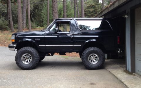1993 Ford Bronco - A black Ford Bronco is parked on a paved surface next to a garage. The vehicle is shown from the side with its front tire turned slightly t…