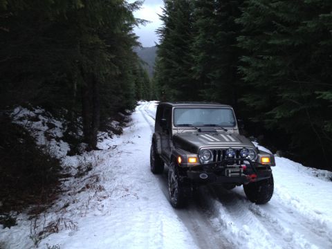 2005 Jeep Wrangler - A silver Jeep Wrangler is driving on a snow-covered dirt road through a dense evergreen forest. The vehicle has aftermarket lights and a wi…
