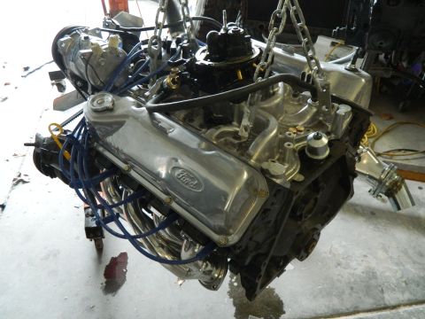 1979 Ford Bronco - A V8 engine with chrome valve covers and exhaust headers is suspended by chains. The engine is partially assembled with blue spark plug wir…