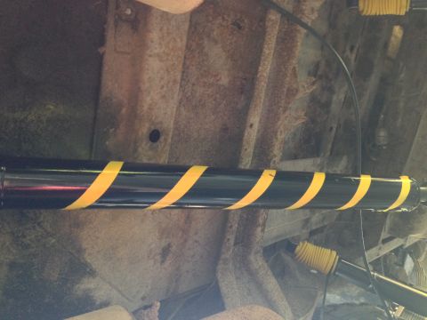 1979 Ford Bronco - The underside of a vehicle shows a black driveshaft with yellow hazard tape wrapped around it in a spiral pattern. The vehicle's rusty fram…