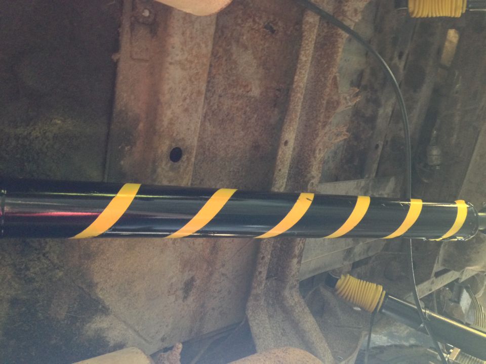 Underside of a vehicle showing a driveshaft with yellow hazard tape and rusty frame.