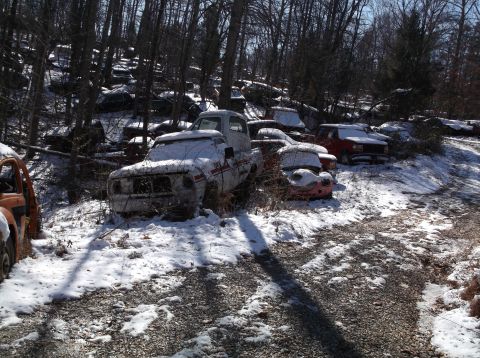 1974 Dodge Challenger - A collection of abandoned vehicles, mostly trucks and cars, are covered in snow in a wooded area. A white pickup truck is prominently featu…