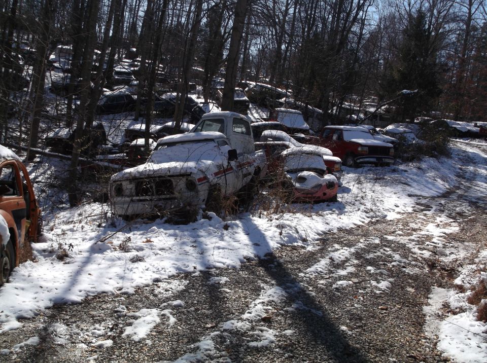 Snow-covered abandoned vehicles in a wooded area, with a white pickup truck in the foreground.